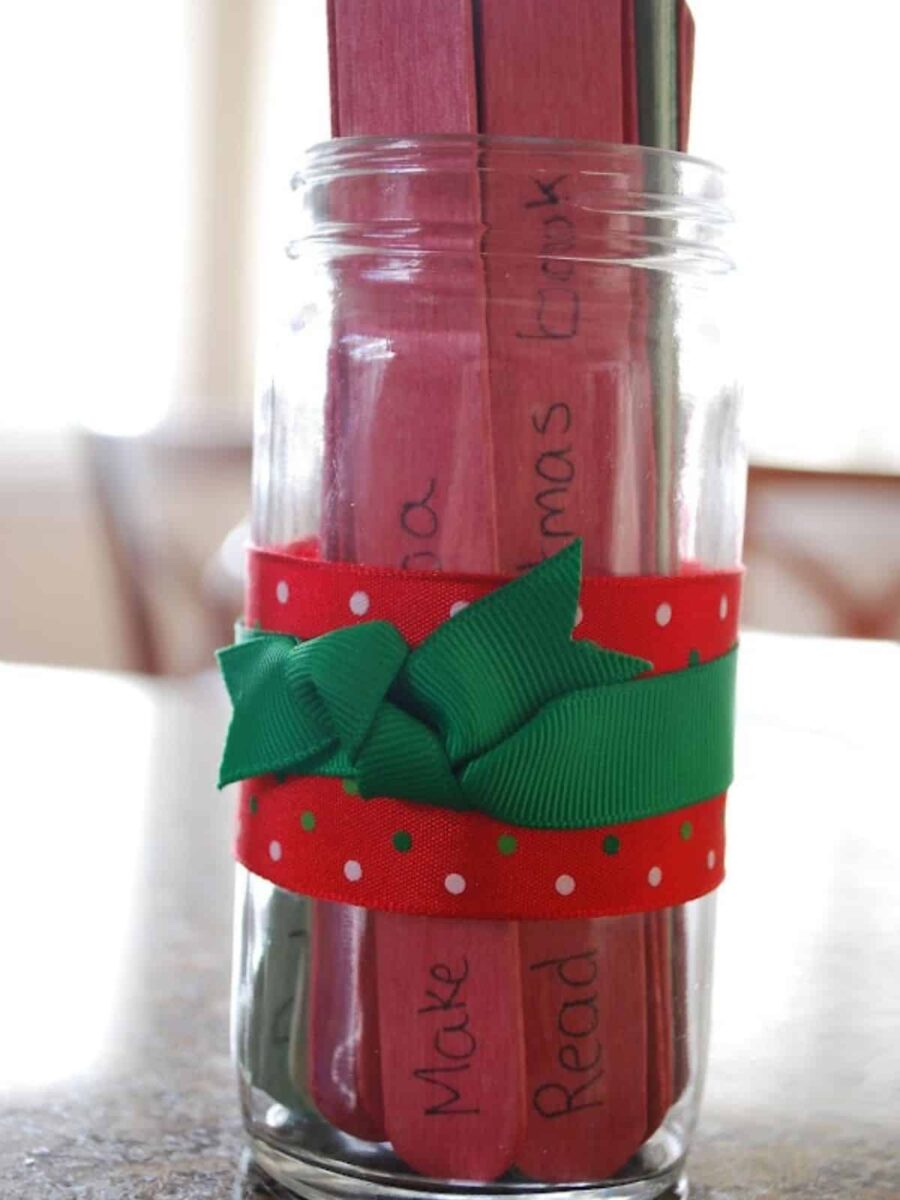 A clear glass jar with red and green painted craft sticks inside, wrapped with a red ribbon with white polka dots and a green bow around the middle. Some sticks have handwritten words visible. advent calendar ideas for adults