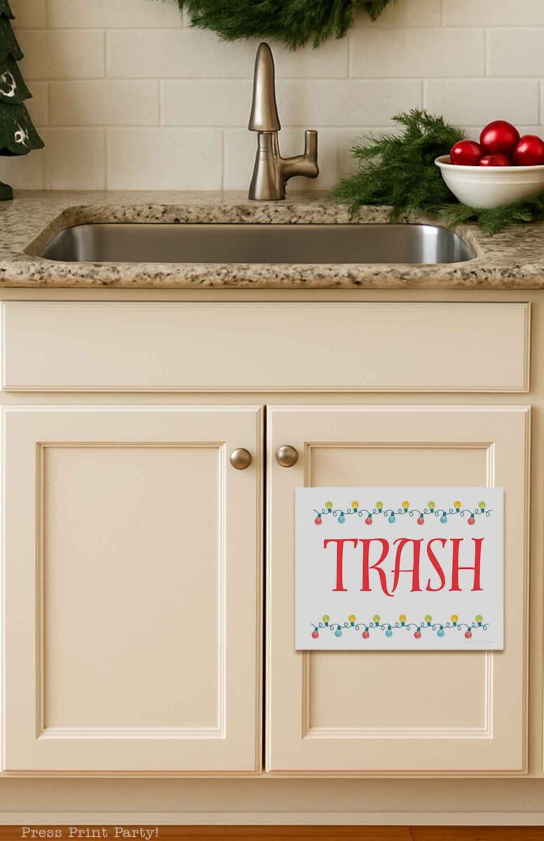A kitchen sink with a granite countertop, festive greenery, and red ornaments nearby. A sign labeled "TRASH" is attached to the cream-colored cabinet below the sink.