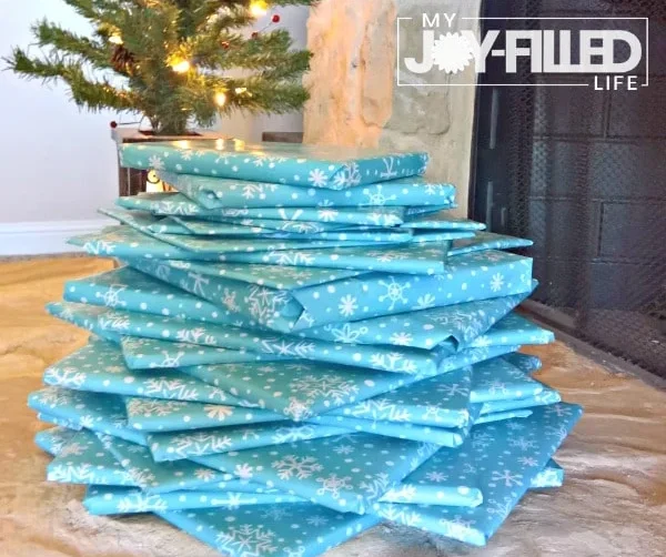 A stack of books wrapped in blue snowflake paper sits by a Christmas tree. Text reads: "Christmas Book Advent Countdown on the Cheap. My Joy-Filled Life.