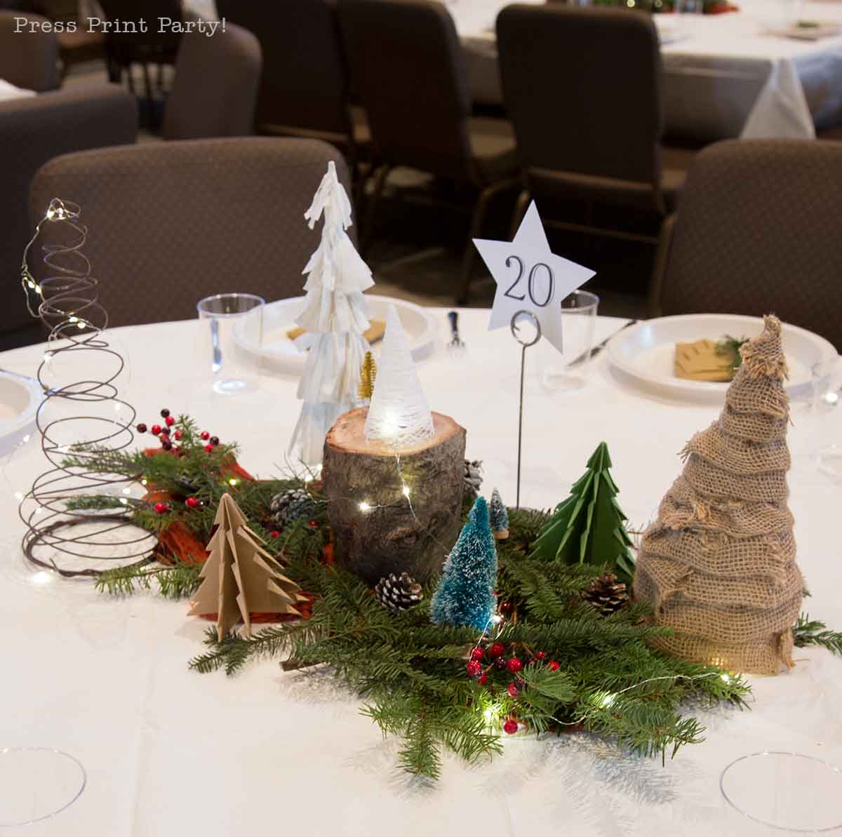 A round table decorated with a festive winter centerpiece featuring small model trees, greenery, berries, a burlap-wrapped tree, a wire tree, fairy lights, and a star-shaped table number 20. Christmas party centerpiece