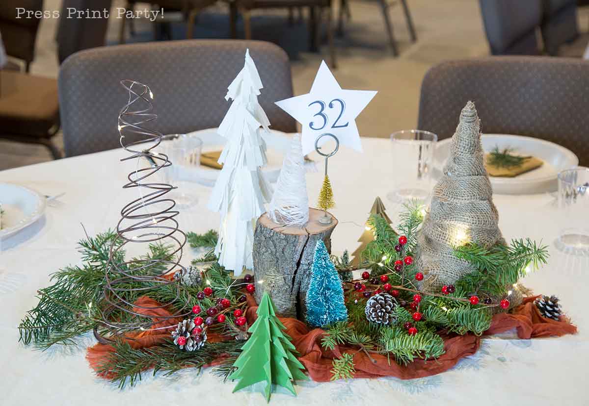 A festive table centerpiece features mini Christmas trees made of paper, twine, and wire, along with pine branches, red berries, pinecones, and a wooden stump holding a star-shaped table marker with the number 32.