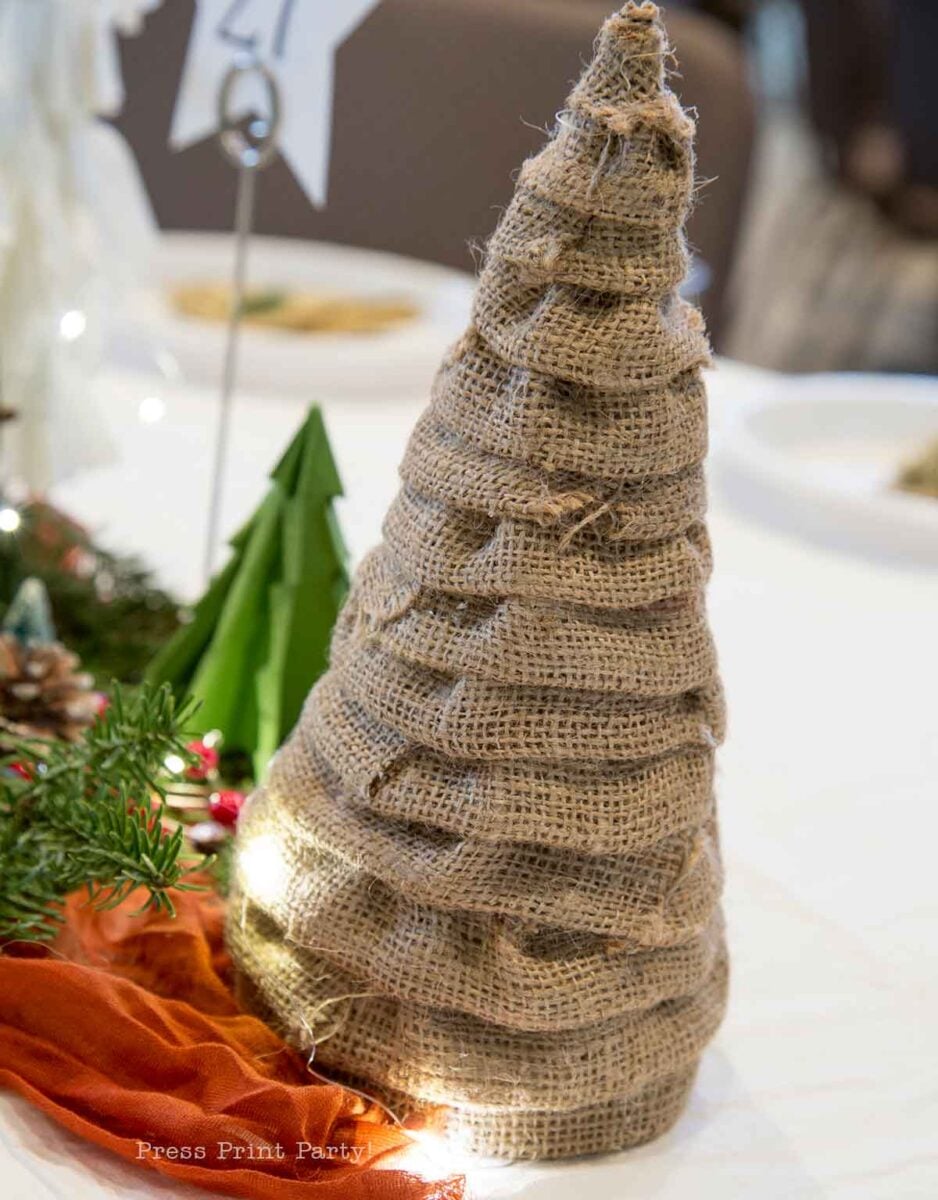 A tabletop decoration shaped like a Christmas tree, made from wrapped burlap fabric, sits on a white table next to greenery, a small green tree, and an orange cloth.
