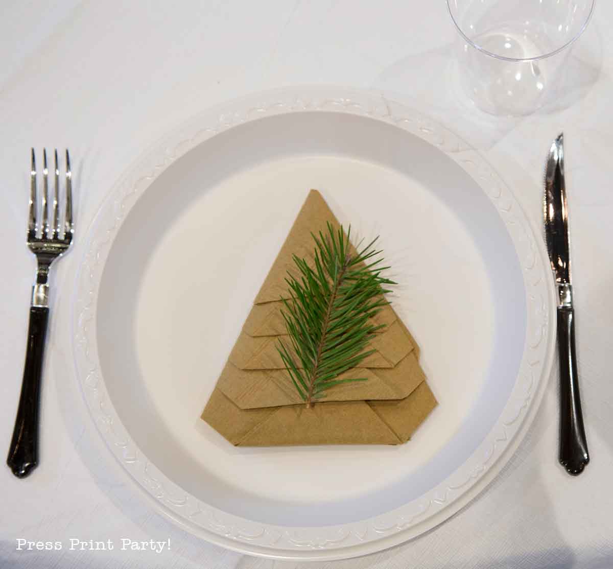 A white plate with a brown napkin folded into a Christmas tree shape, topped with a small pine branch. The plate is set with a fork, knife, and clear plastic cup on a white tablecloth. Christmas party centerpiece