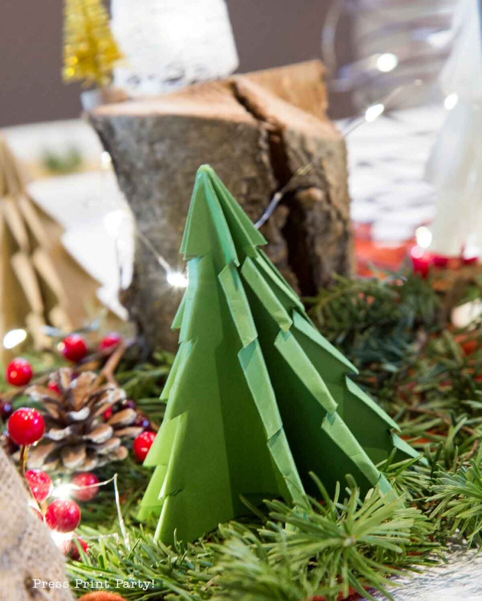 A green paper Christmas tree centerpiece sits on a table decorated with pine branches, red berries, a pine cone, and logs, with fairy lights in the background.