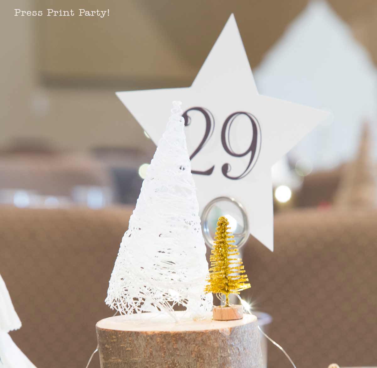 A white string Christmas tree and a small gold tree sit on a wooden base in front of a white star with the number 29, used as a table centerpiece at an event.