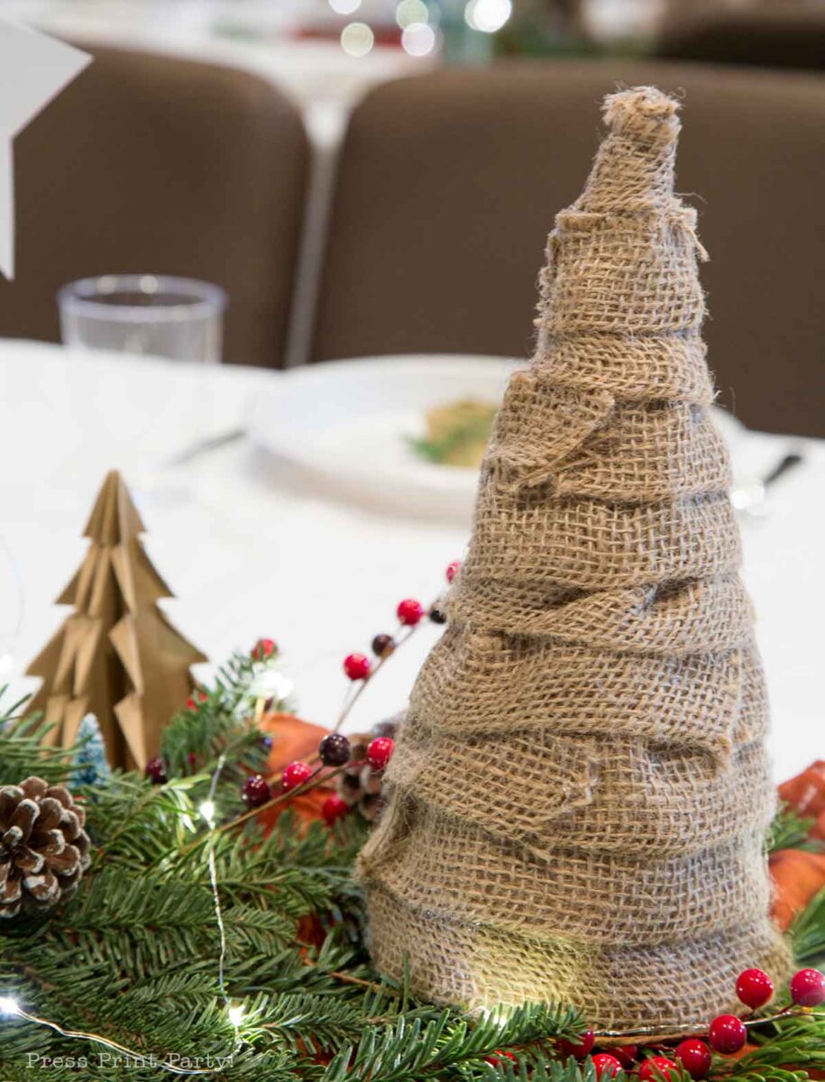A burlap-wrapped cone shaped like a Christmas tree sits on a festive table, surrounded by greenery, red berries, pinecones, and a small wooden tree decoration. Christmas party centerpiece