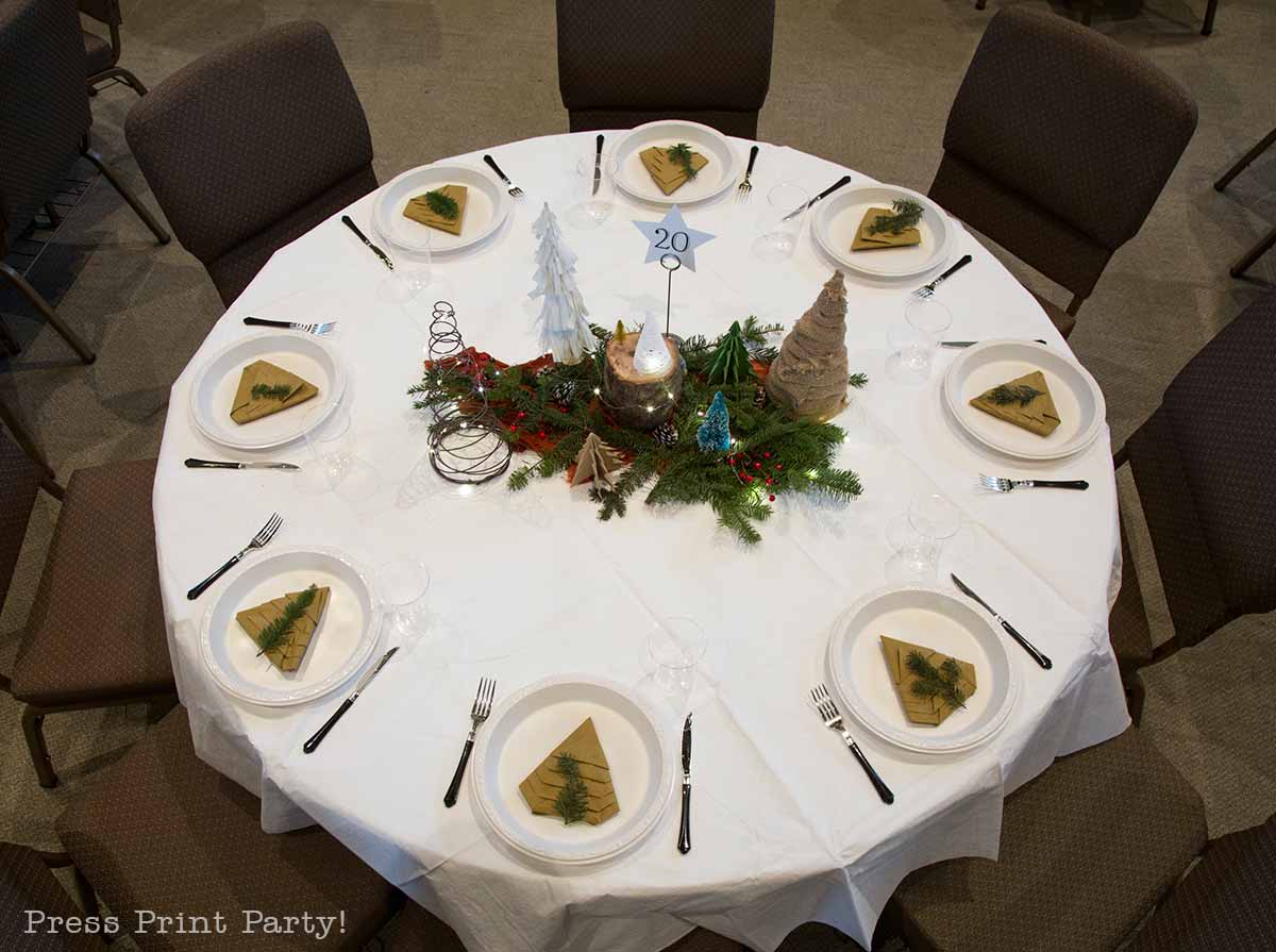 A round table set for eight with white plates, napkins folded as Christmas trees, cutlery, wine glasses, and a festive centerpiece featuring greenery, candles, and decorative trees. A table number “20” is displayed. Christmas party centerpiece