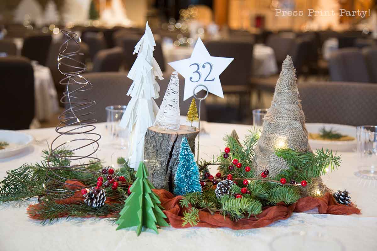 Table centerpiece with pine branches, red berries, pinecones, miniature trees made of various materials, a tree stump, and a star-shaped table number 32, set on a white tablecloth in a banquet setting.