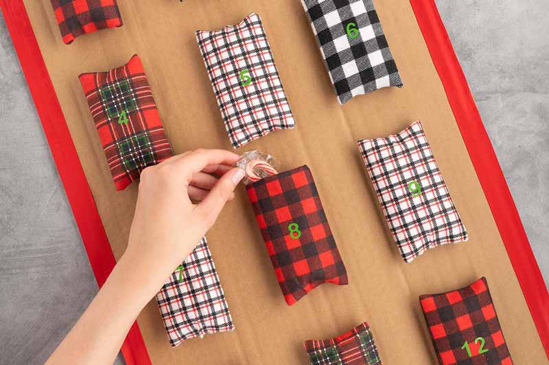 A hand places a candy cane into a black and red plaid pouch labeled with a green "8," among several plaid pouches numbered for an advent calendar on a brown background.