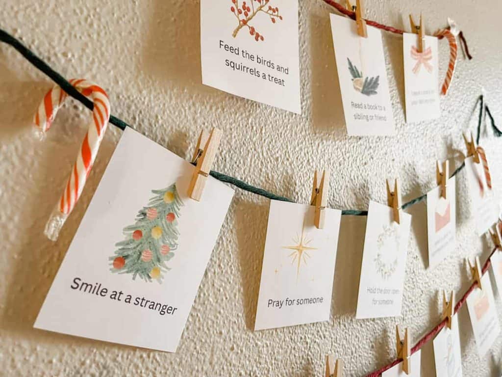 Holiday-themed cards with positive messages, such as "Smile at a stranger" and "Pray for someone," hang from twine on a wall using small clothespins. A candy cane also hangs with the cards. advent calendar ideas for adults and teens