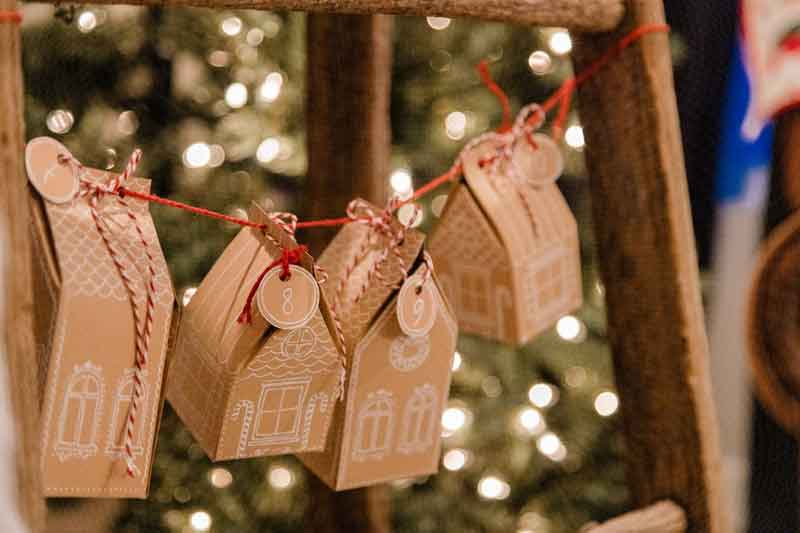 Small cardboard houses with numbered tags hang on red string, likely as part of an advent calendar, against a blurred background of Christmas tree lights and decorations. advent calendar ideas for adults