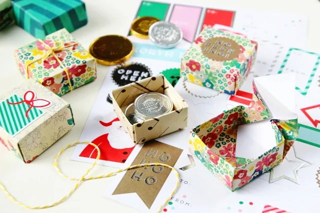 Small, colorful paper gift boxes, some open and some closed, filled with chocolate coins, arranged on a table with festive tags, string, and wrapping materials in the background.