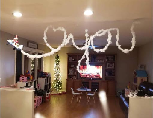 A living room decorated for Christmas features cotton โcloudsโ hanging from the ceiling, spelling out โI โค๏ธ you.โ Thereโs a decorated Christmas tree, toys, a small table, and holiday lights in the background.
