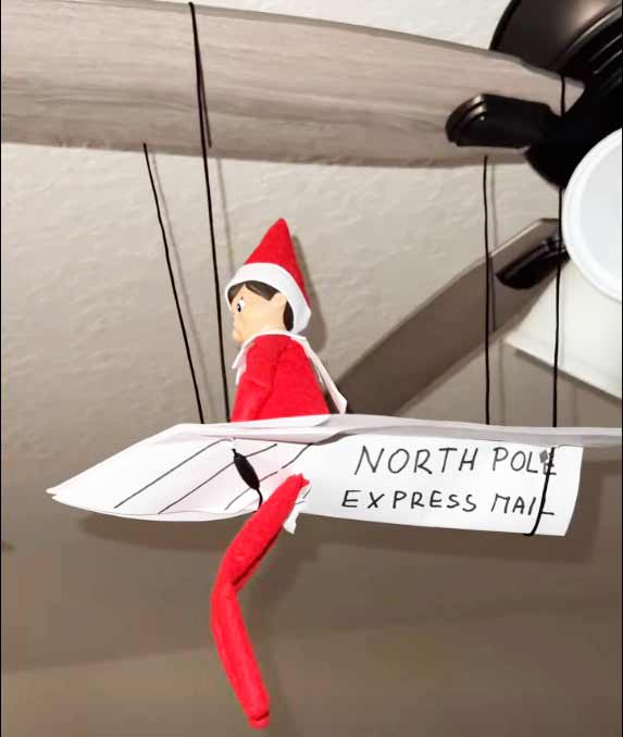 A toy elf dressed in red sits on a paper airplane labeled "North Pole Express Mail," suspended from the blades of a ceiling fan.