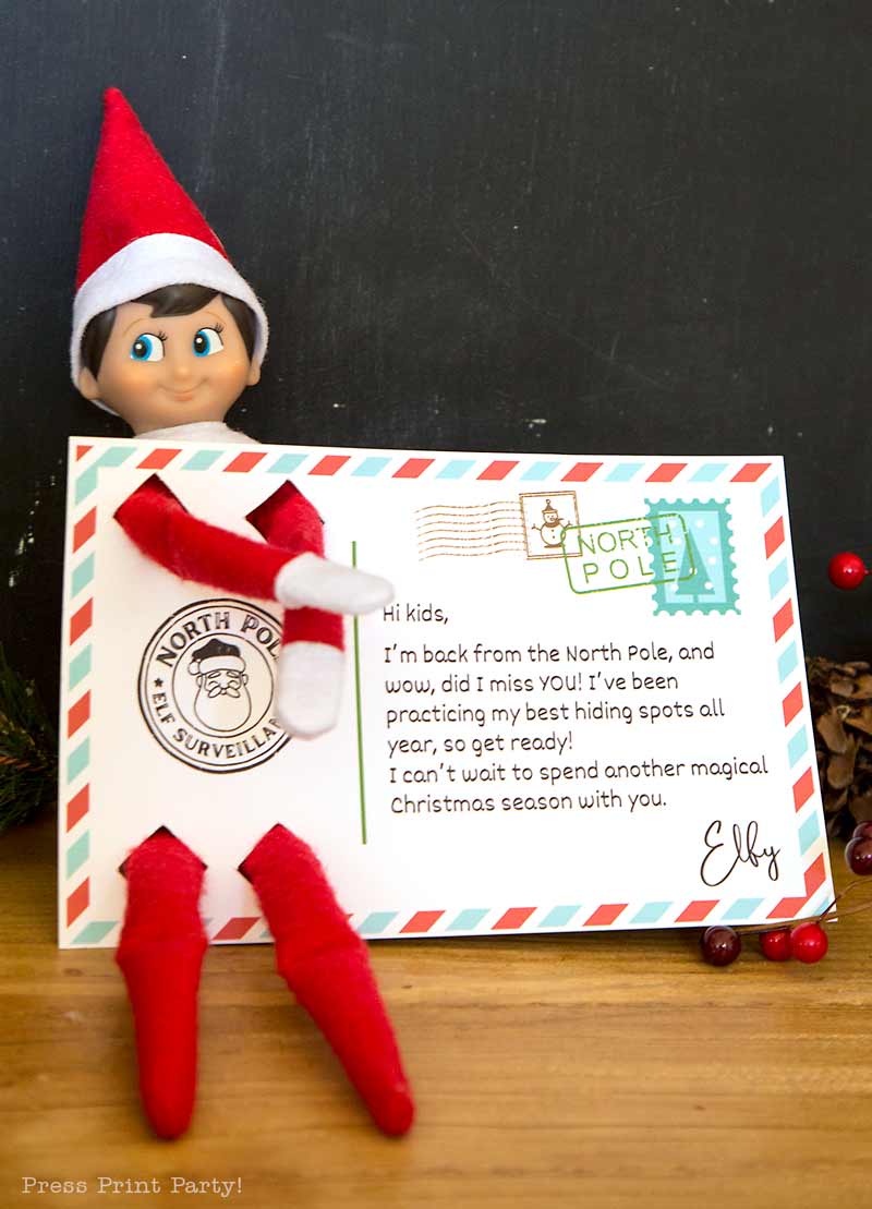 A toy elf holds a postcard featuring North Pole-themed stamps and a message for kids, sitting on a wooden surface with holiday decor in the background.