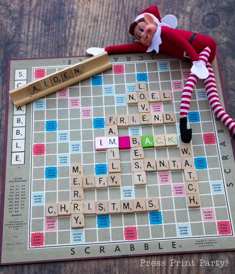 A Scrabble board spells out "LOVE, FAMILY, I'M BACK, MERRY CHRISTMAS," with an Elf on the Shelf doll lying on the board and wooden tiles spelling "AIDEN" above. The board sits on a wooden table.