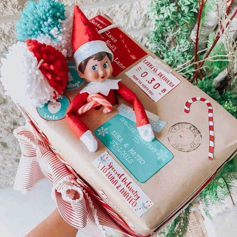 A Christmas gift wrapped in brown paper with colorful labels, a red and white candy cane sticker, and a small plush Elf on the Shelf doll sitting on top. Red, green, and white pom-poms decorate the package.
