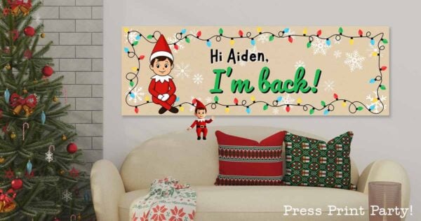 A festive living room with a decorated Christmas tree and a sofa with holiday pillows. Above the sofa is a banner with an elf, colorful lights, snowflakes, and the text "Hi Aiden, I'm back!.