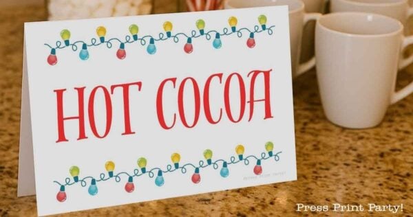 A festive "Hot Cocoa" sign decorated with colorful string lights sits on a countertop near several white mugs.