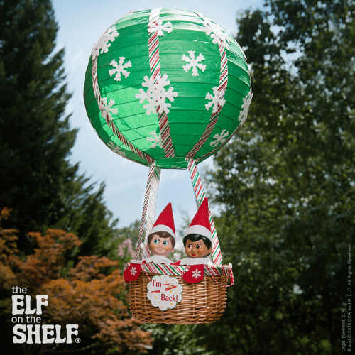 Two Elf on the Shelf dolls sit in a basket attached to a green hot air balloon decorated with white snowflakes and red stripes. The basket has a sign that reads "I'm Back!" Trees and sky are in the background.