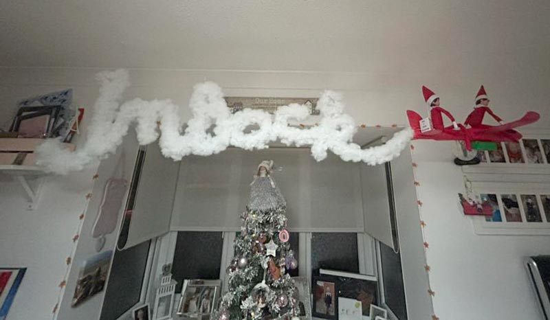 A living room decorated for Christmas features a Christmas tree, a "Indeck" sign made of fluffy material above a window, and two elf dolls riding a red airplane hanging from the ceilingโperfect for creative elf on the shelf arrival ideas.