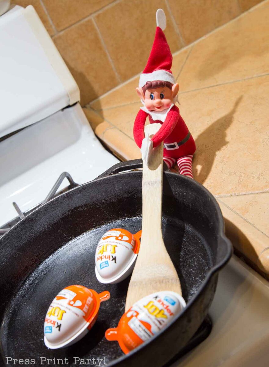 A toy elf holds a large wooden spoon, stirring three Kinder Joy eggs in a black cast iron skillet on a stovetopโa playful scene perfect for simple elf shelf ideas. The stove and tiled countertop are visible in the background.