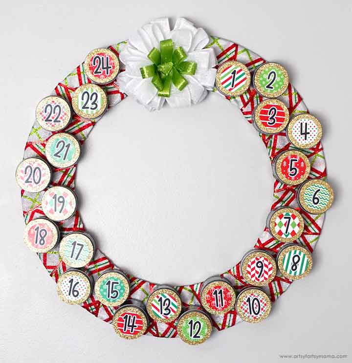 A circular Christmas advent calendar wreath with 24 numbered sections, decorated in red, green, and white patterns, and topped with a large white and green bow at the top.