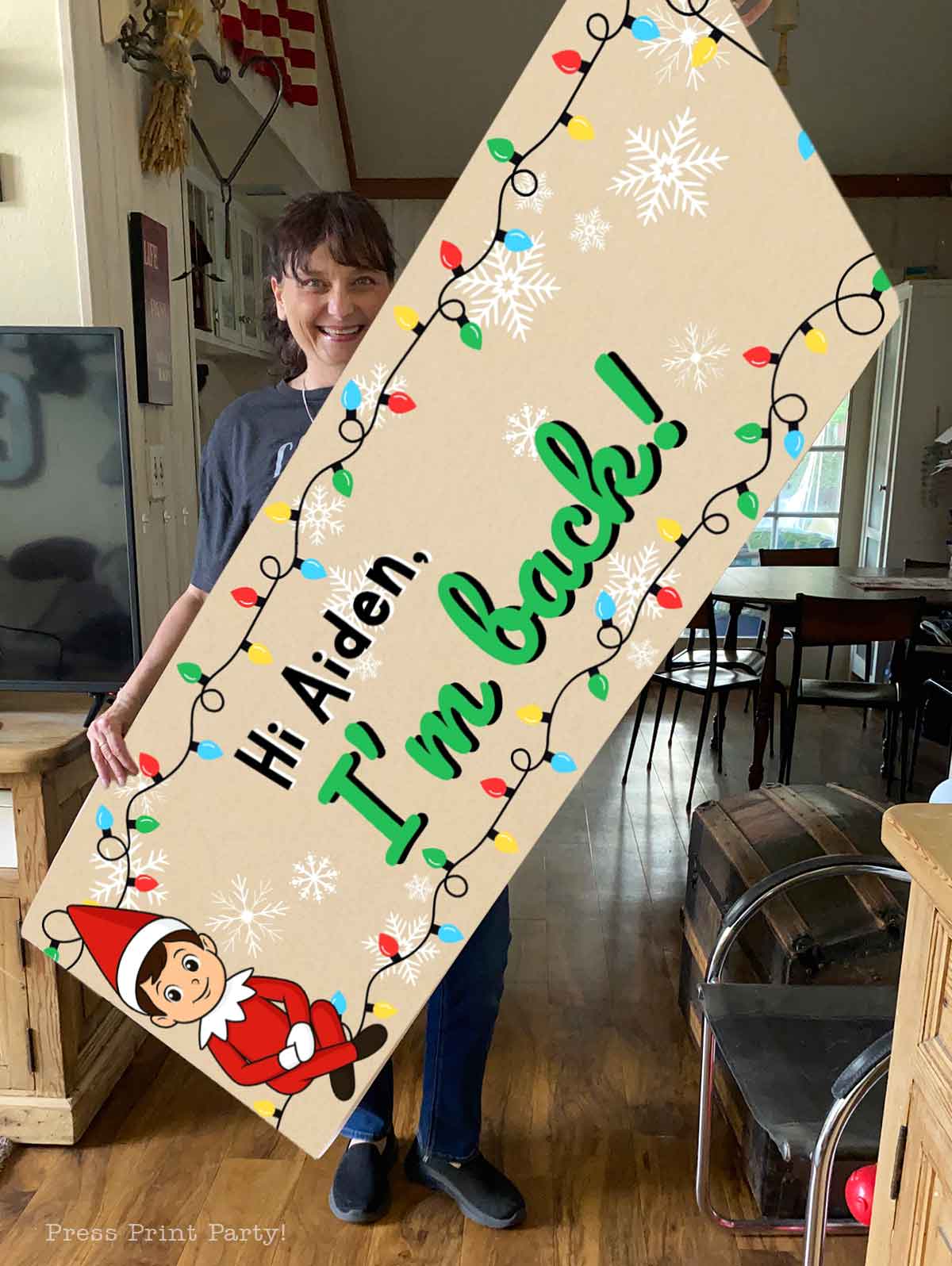 A smiling woman holds a large, festive elf on the shelf sign decorated with snowflakes and string lights that reads, "Hi Aiden, I'm back!" with a cartoon elf in the corner.