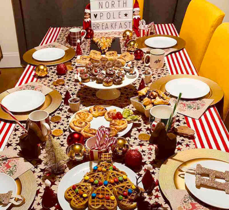 A festive table is set with Christmas-themed treats, mugs, and decorations. A sign reads "NORTH POLE BREAKFAST." The tablecloth is red and white striped, and there are ornaments and holiday shapes throughout.