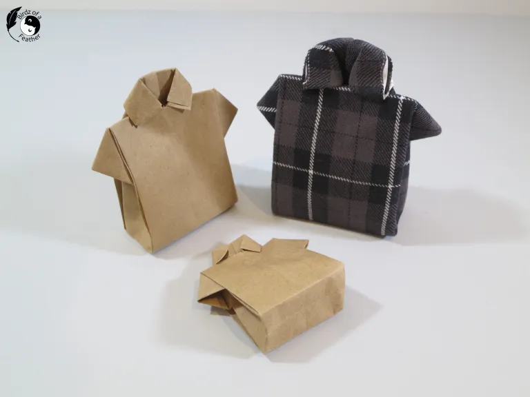 Three shirt-shaped gift boxes are displayed on a white surface. Two are made from brown paper and one is wrapped in black-and-gray plaid fabric. Each has a folded collar and short sleeves.