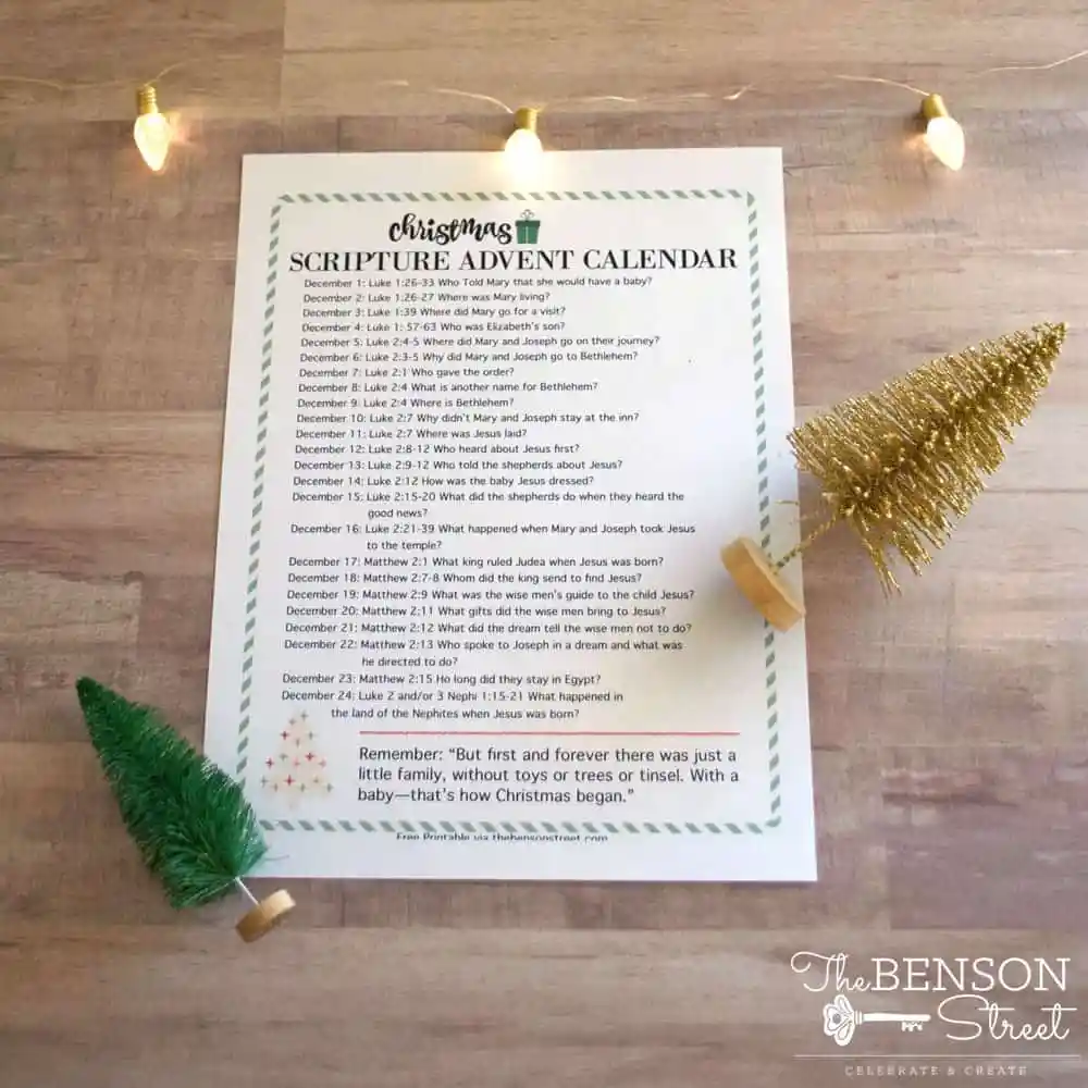 A Christmas scripture advent calendar is displayed on a wooden surface with two small Christmas trees and string lights. The calendar lists Bible readings for December days. "The Benson Street" is in the bottom right corner.