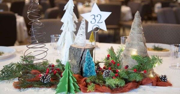 A festive table centerpiece features charming christmas tree table decorations made of wood, burlap, and wire, surrounded by pine branches, berries, pinecones, and a table number 32 on a star-shaped card.