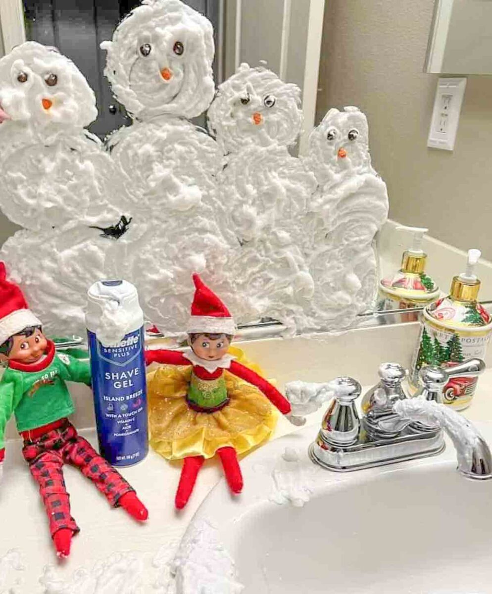 Two elf dolls sit on a bathroom sink in front of a mirror covered in shaving cream snowmen. A can of shave gel and soap bottles are also on the counter. The scene looks playful and festive.