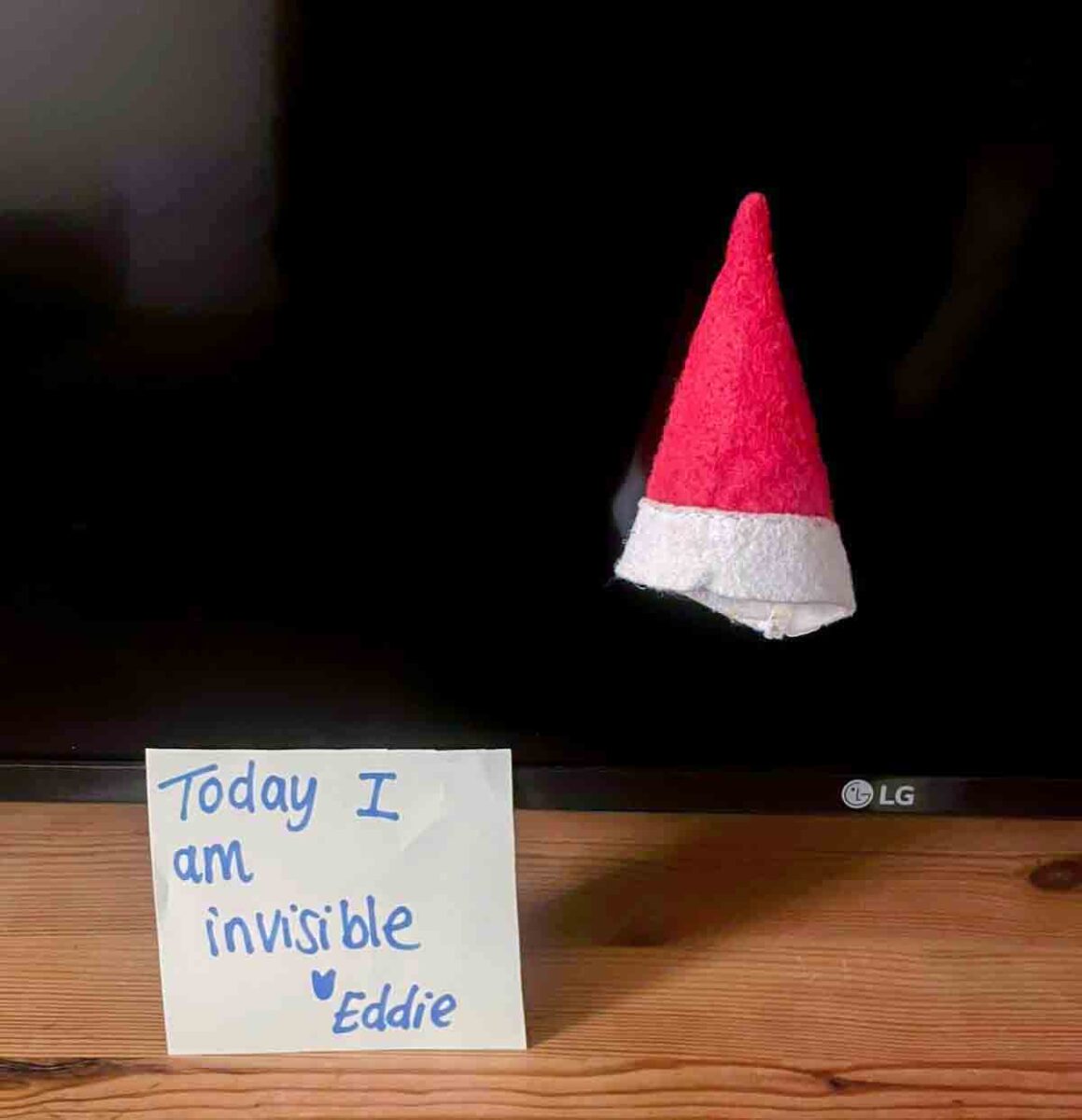 A red and white Santa hat appears to float in front of a TV screen. Below it, a note reads, "Today I am invisible โฅ Eddie," suggesting someone is pretending to be invisible except for the hat.