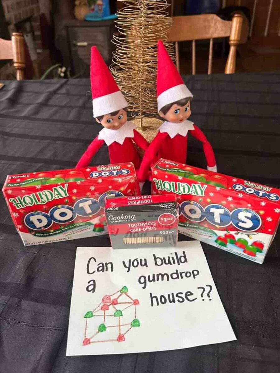 Two Elf on the Shelf dolls sit behind boxes of Holiday Dots candy and a box of toothpicks. In front is a note that reads, "Can you build a gumdrop house??" with a drawing of a house built from candy.