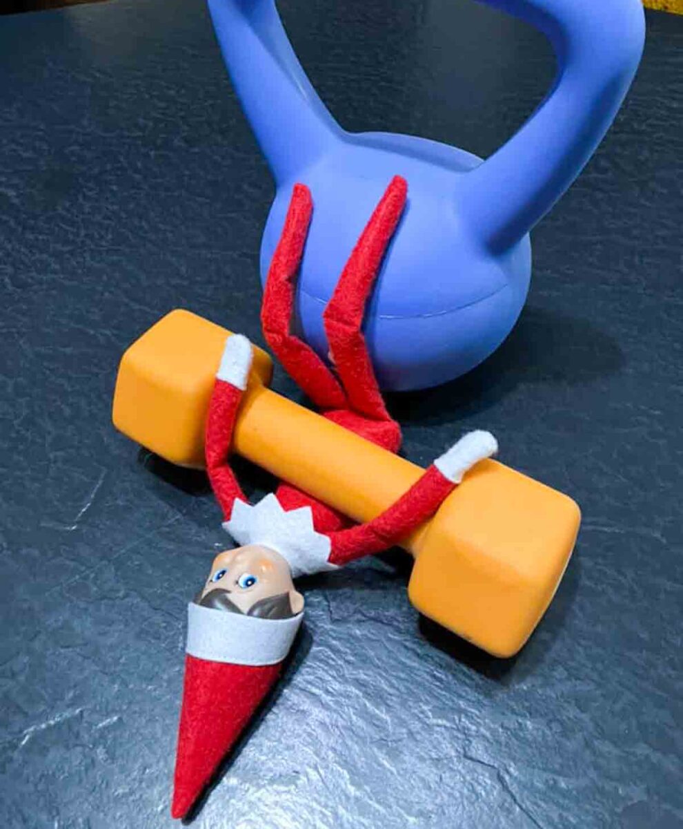 A toy elf with red and white clothing is positioned upside down, holding a yellow dumbbell, with its legs resting against a blue kettlebell on a dark textured surface.