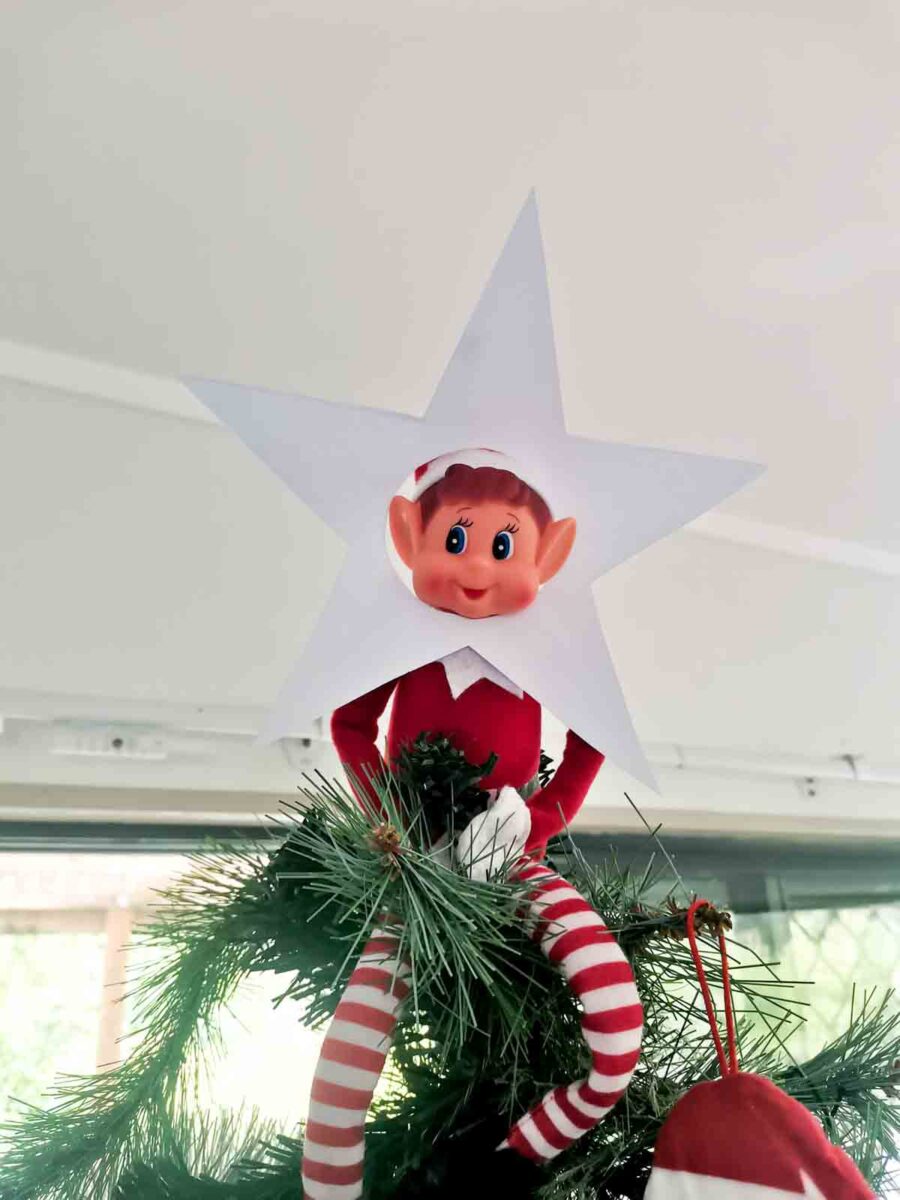 A festive elf doll with a paper star around its face sits atop a Christmas tree, surrounded by green pine branches and red and white striped legs dangling down.