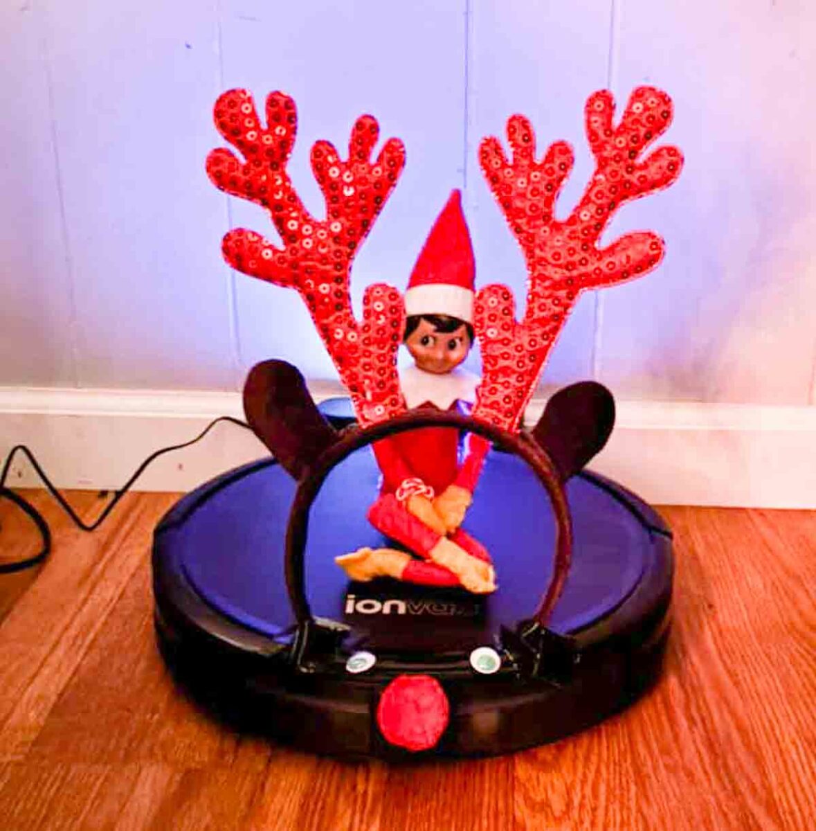 An Elf on the Shelf doll sits on a robotic vacuum cleaner that has red, sparkly antlers attached, resembling a reindeer, on a wooden floor against a light-colored wall.