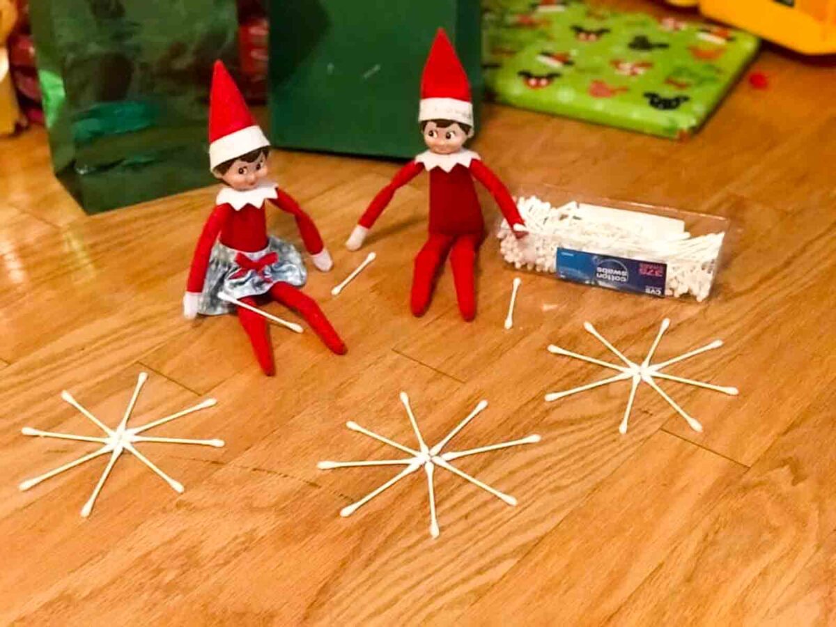 Two Elf on the Shelf dolls sit on a wooden floor next to a box of cotton swabs, which are arranged in the shape of snowflakes in front of them. Bright toys are visible in the background.