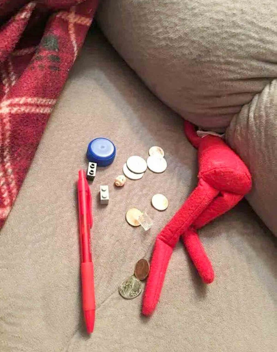 A red pen, assorted coins, dice, a blue bottle cap, small beads, and a red plush toy are scattered on a gray sofa next to a red plaid blanket.