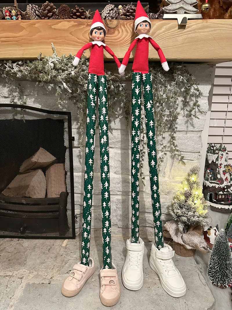 Two Elf on the Shelf dolls with unusually long legs wearing green pants with white trees stand side by side in shoes, posed in front of a decorated fireplace and Christmas decorations.