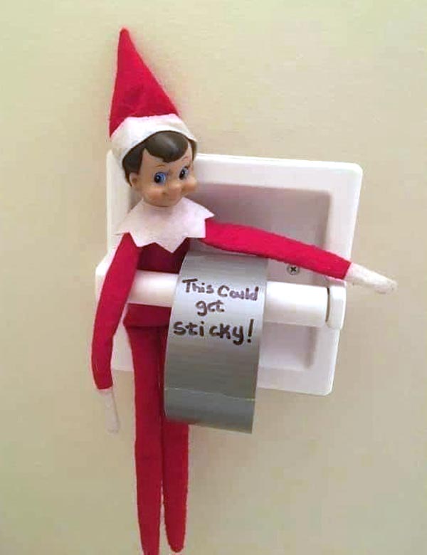 A toy elf sits on a toilet paper holder with a roll wrapped in silver duct tape. The tape is labeled "This could get sticky!" in black marker. The elf appears to be holding the roll.