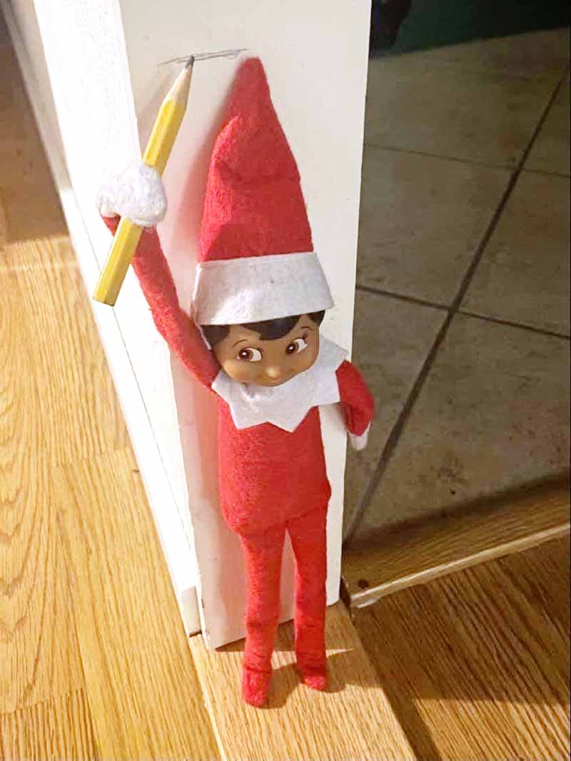 A toy elf in a red outfit stands against a white wall, holding a yellow pencil and marking a line above its head, as if measuring its height. Hardwood and tile flooring are visible.