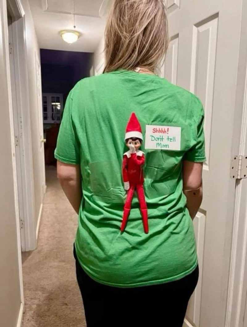 A person with long blonde hair wears a green shirt with an Elf on the Shelf doll attached to the back. The elf holds a sign that says "Shhh! Don't tell Mom." The scene is in a hallway with doors on both sides.