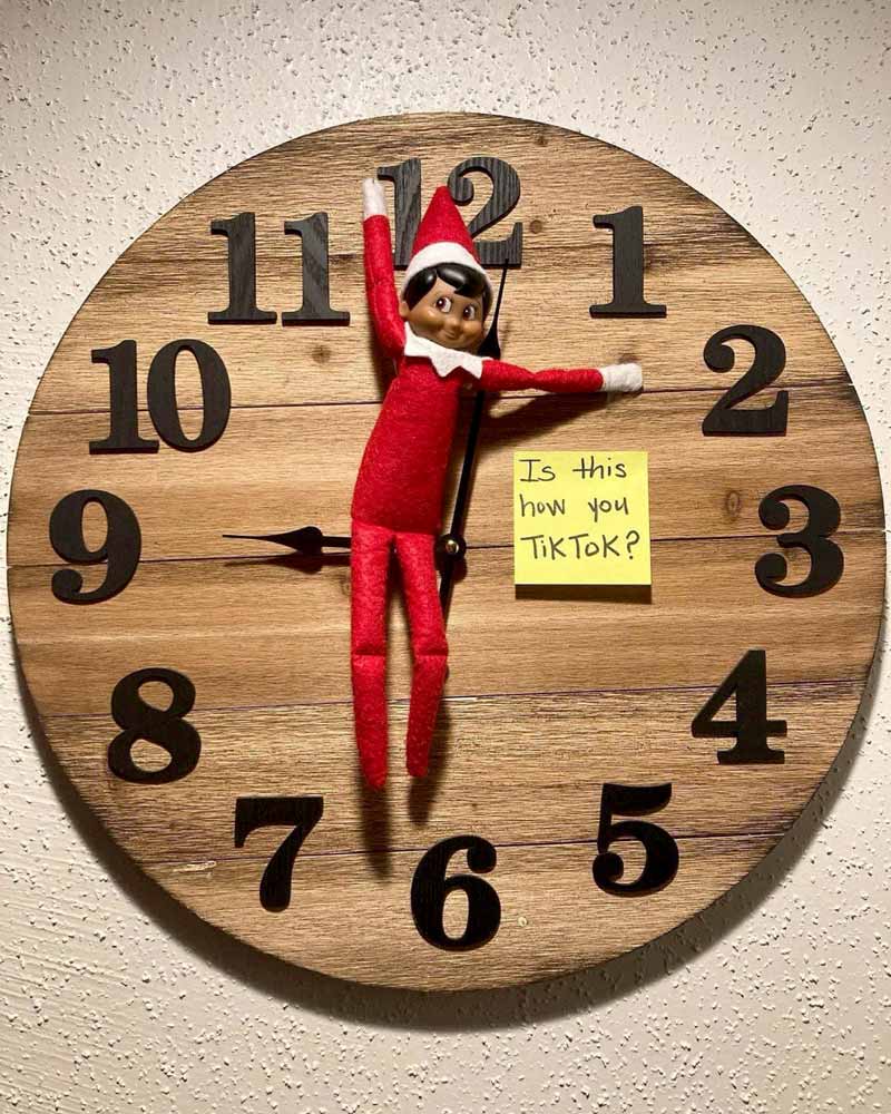 A wooden wall clock shows the time as 9:00. An Elf on the Shelf doll is pinned between the clock hands, and a yellow sticky note reads, "Is this how you TikTok?.