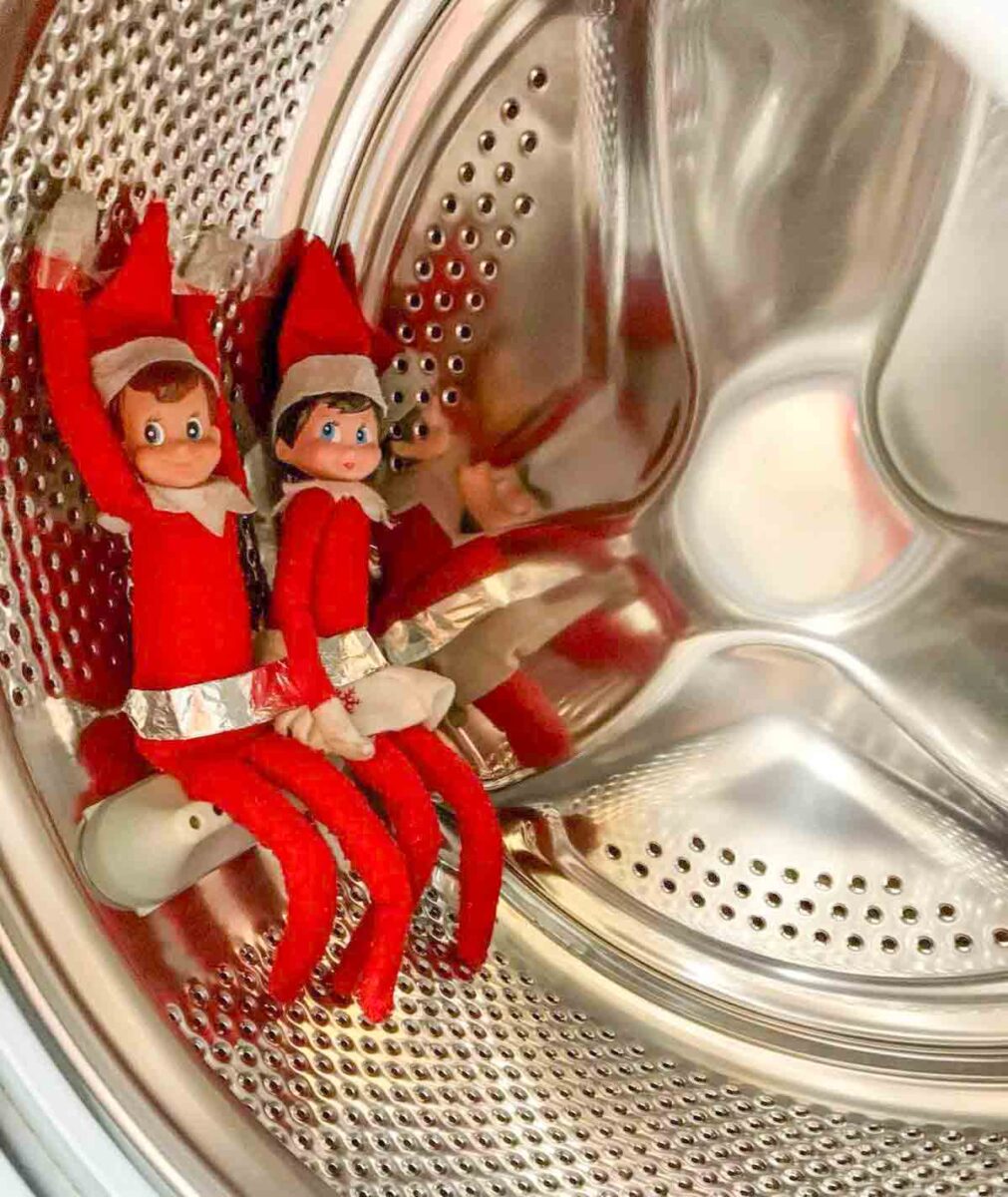 Two toy elves dressed in red outfits with white trim are sitting inside a shiny, empty washing machine drum, appearing as if they are about to go for a spin.