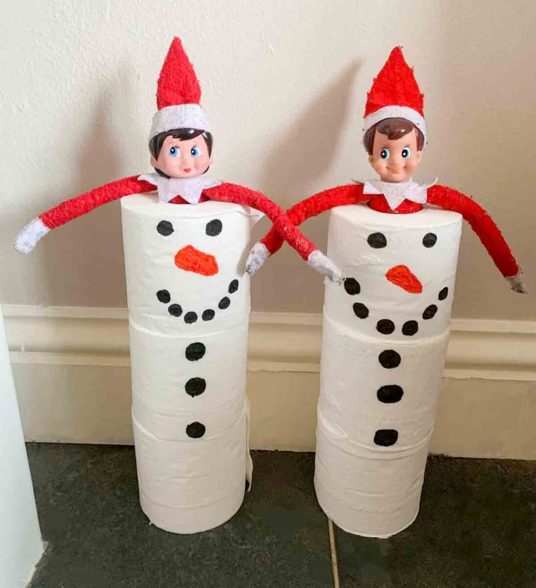 Two elf dolls dressed in red sit on top of stacked toilet paper rolls decorated as snowmen with black marker eyes, buttons, and orange carrot noses. The figures are placed against a white wall on a tiled floor.