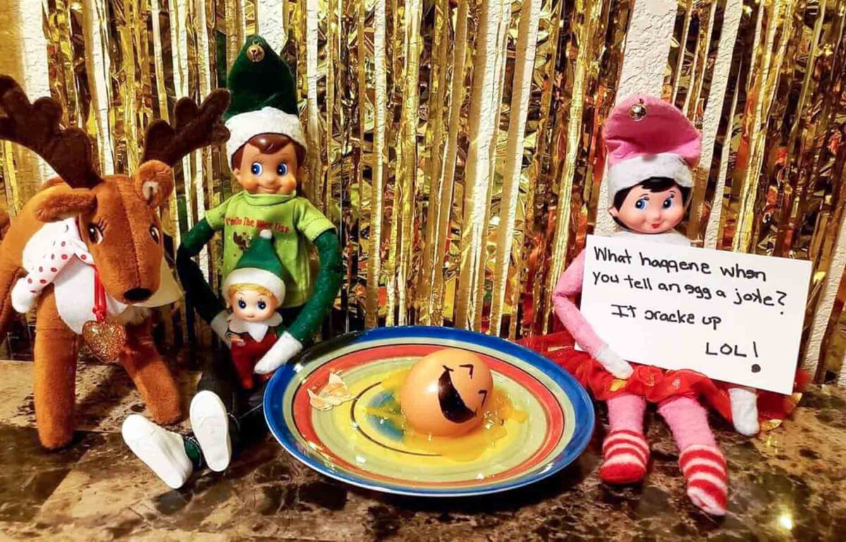 A reindeer plush and three elf dolls sit in front of gold tinsel. One elf holds a sign with a joke. On a colorful plate is a cracked egg with a drawn smiley face. The sign reads: "What happens when you tell an egg a joke? It cracks up LOL!.