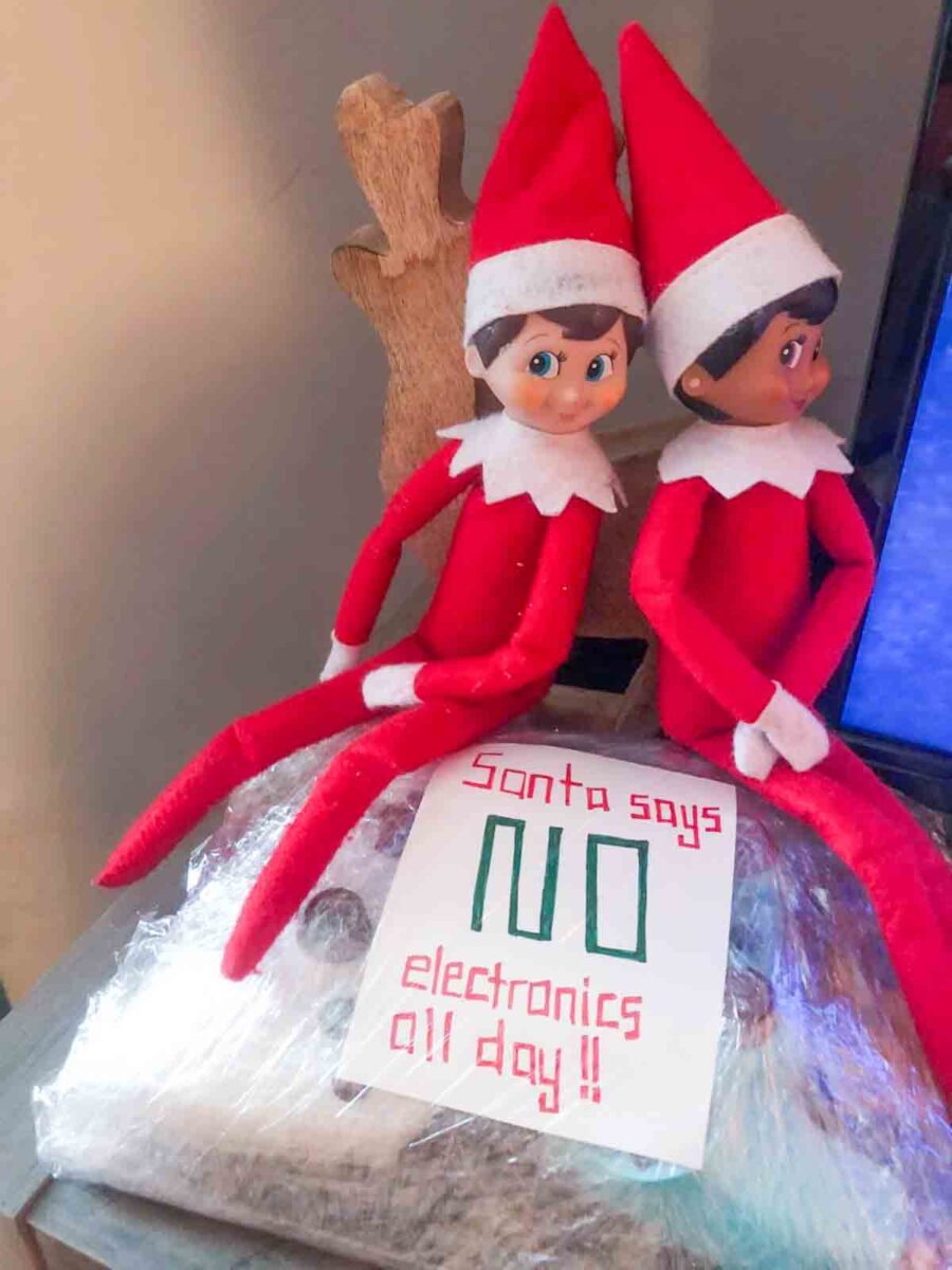 Two Elf on the Shelf dolls sit on a counter next to a reindeer decoration, with a handwritten sign in front that says โSanta says NO electronics all day!!โ and electronics wrapped in plastic wrap underneath.