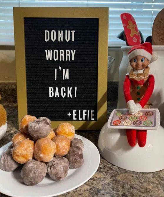 A sign reads "DONUT WORRY I'M BACK! +ELFIE" next to a plate of assorted donut holes and a small elf doll holding a tray of cookies, sitting on a counter.